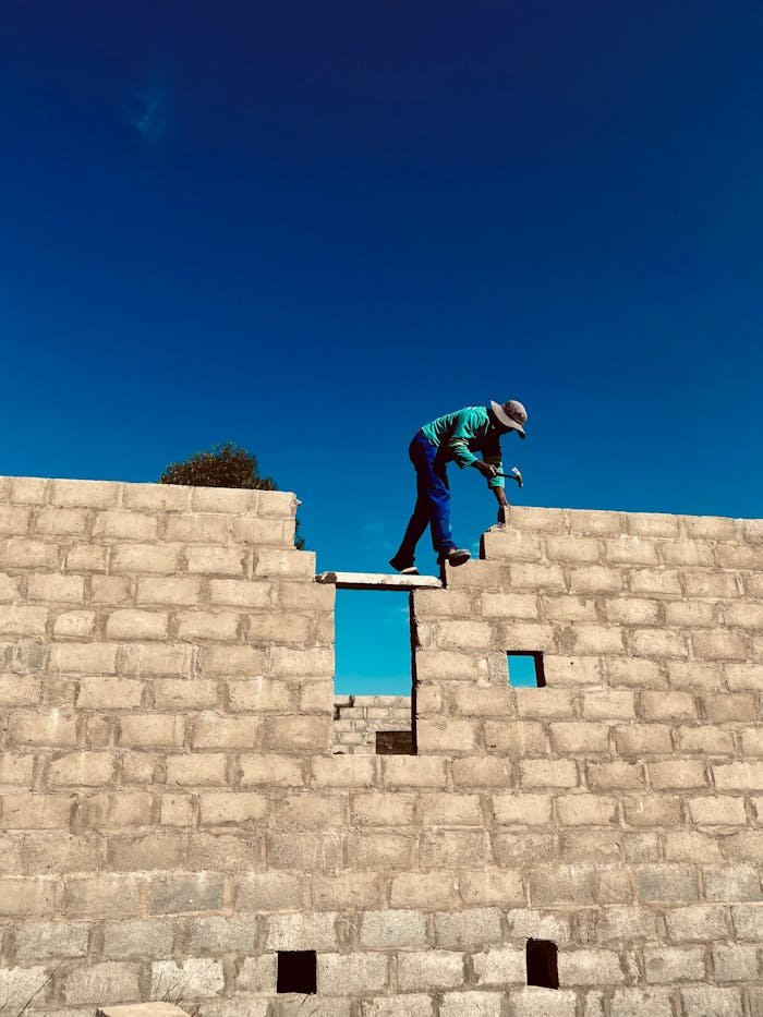 A construction worker balances atop a brick wall with a clear blue sky backdrop.
