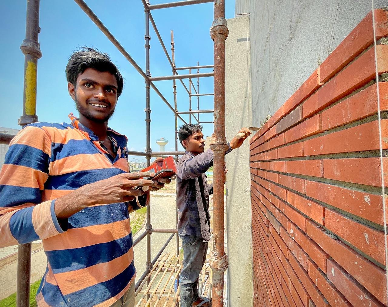 Two construction workers laying red bricks on scaffolding outside a building.